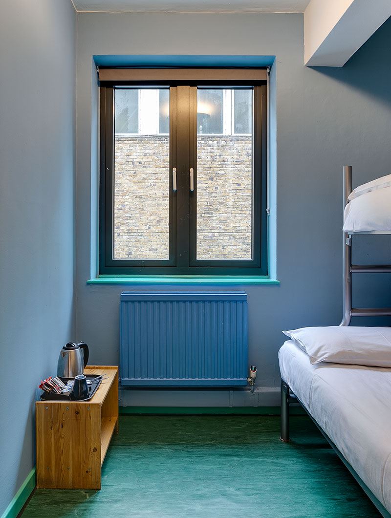 A private room at Clink 261 hostel in London with a double bed and single bed bunk, window, radiator, and shelf with teas, coffee and a kettle.